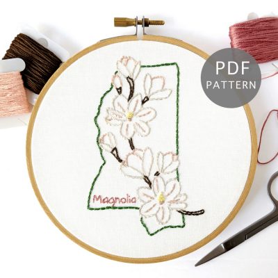 A branch filled with Magnolia flowers stitched on white fabric inside the Mississippi state outline.