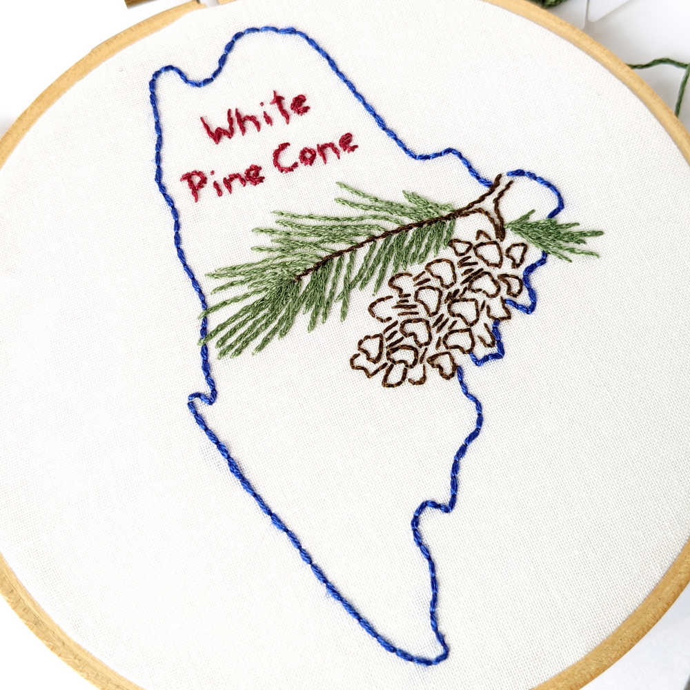 Branch of a white pine cone stitched on white fabric inside the Maine state outline.