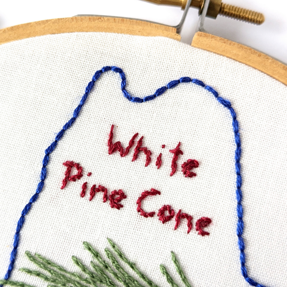 White Pine Cone text stitched in red on white fabric.