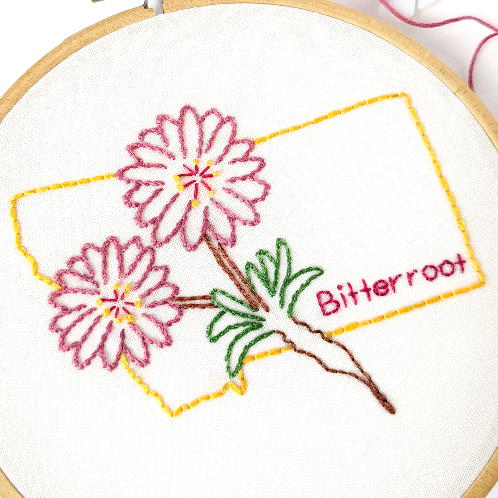 Two Bitterroot flowers stitch on white fabric inside the Montana state outline.