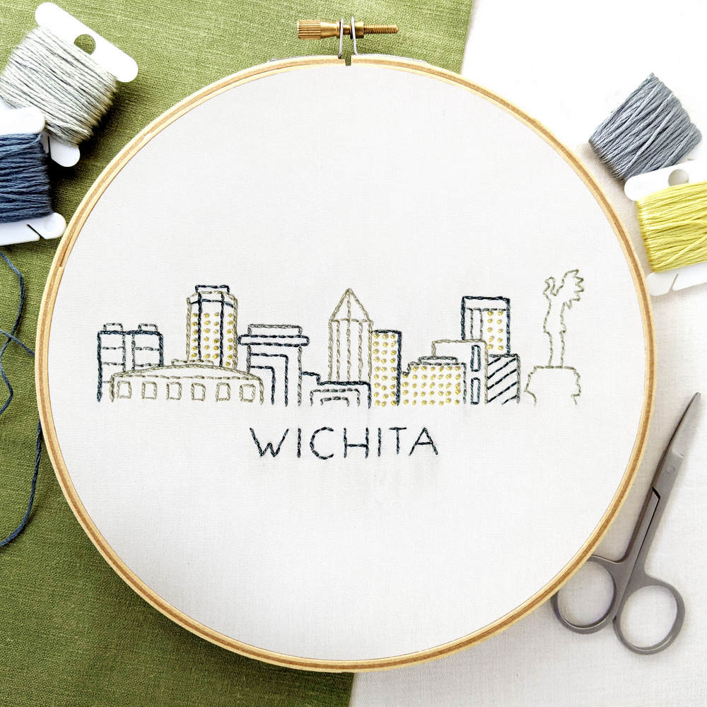 Wichita skyline hand stitched on white fabric inside a wooden hoop. The background is white & green fabric with colored threads and small scissors.