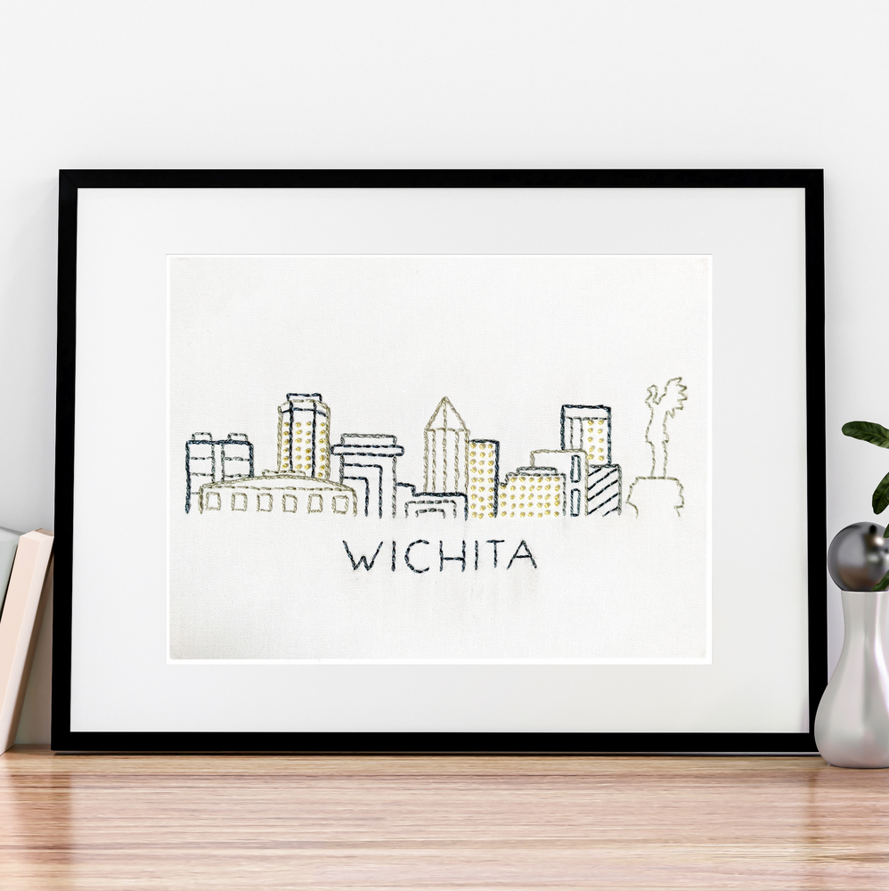 Hand stitched Wichita skyline on white fabric inside a rectangle black frame.